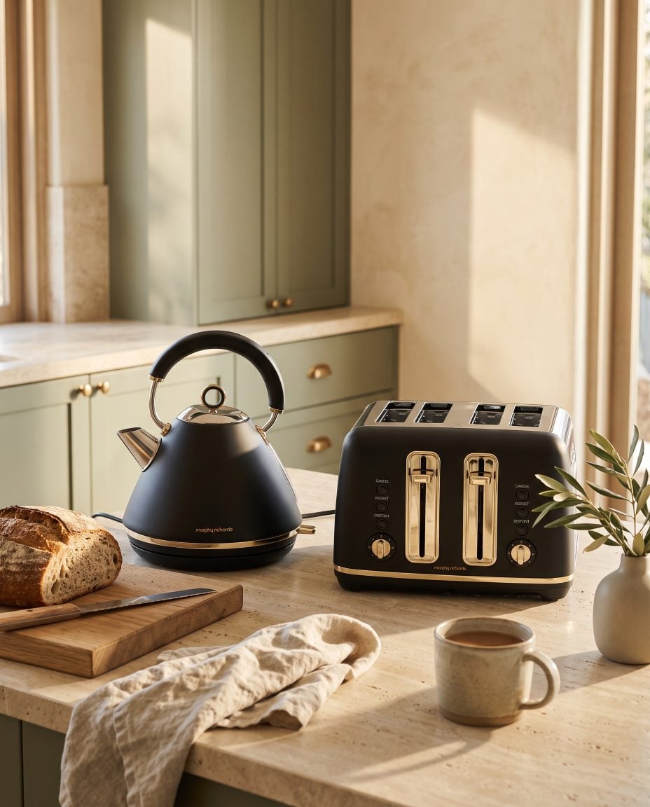 A matte black and soft gold kettle and toaster on a travertine bench in a sage green kitchen with morning light
